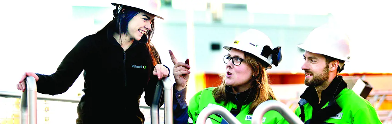 Three people wearing helmets and Valmet jackets engaged in conversation.
