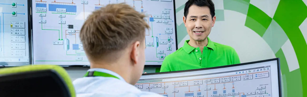 Two Valmet employees in a control room environment reviewing process diagrams on multiple monitors, with one person smiling and engaging in conversation.