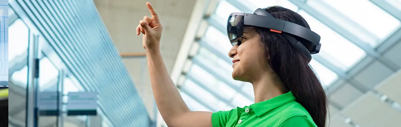A woman wearing a green Valmet shirt and a mixed reality headset interacts with a virtual interface using hand gestures, inside a modern, glass-roofed facility.