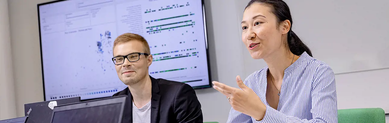 Two colleagues in a meeting room discussing data visualizations displayed on a large screen, with laptops in front of them.