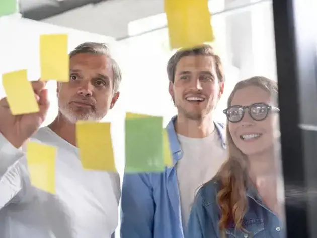 Three coworkers collaborating in an office using sticky notes on a glass wall.
