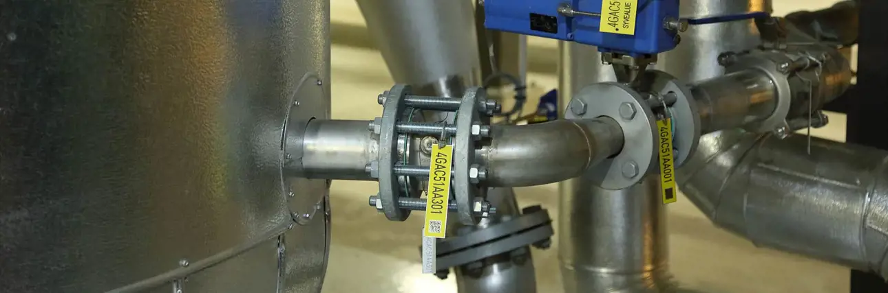 Close-up view of industrial metal piping connected to a large cylindrical tank, featuring bolted flanges, yellow identification tags, and a blue sensor or valve mounted on the pipe.