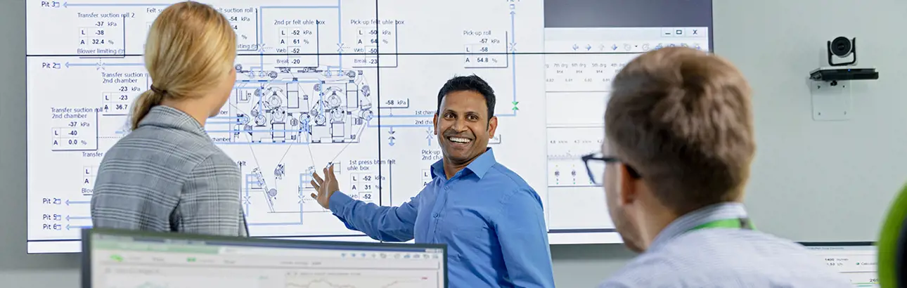 A smiling presenter in a blue shirt leads a technical discussion in front of a large screen displaying a detailed industrial diagram, while two colleagues listen attentively.
