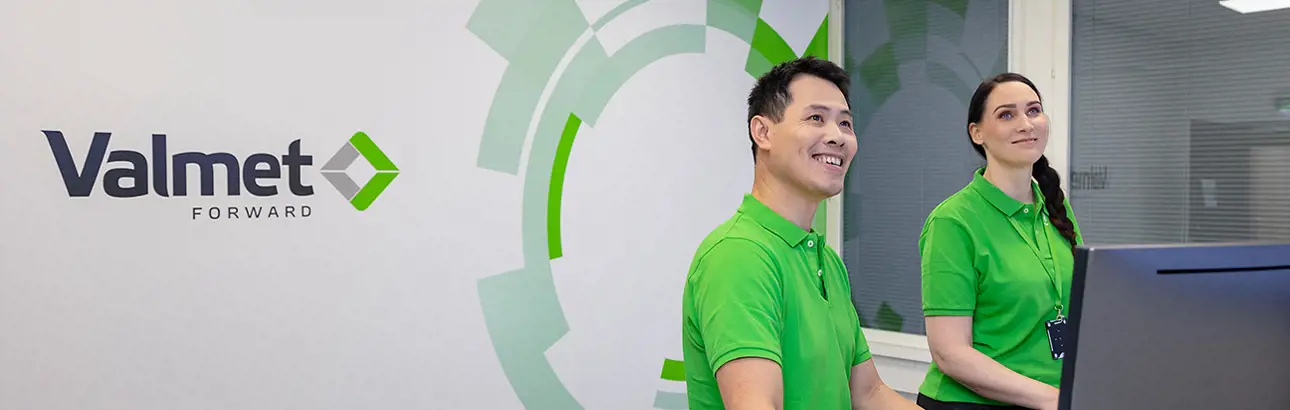 Two smiling Valmet employees in bright green uniforms standing at a reception or information desk, with the Valmet logo and the word 'Forward' displayed on the wall behind them.