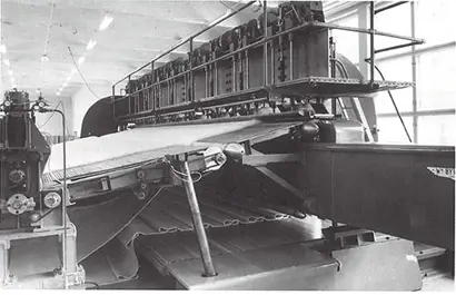 A black-and-white photograph shows a large needling machine in operation inside a well-lit factory. The equipment features multiple rollers and a wide sheet of paper being processed through the system. Elevated platforms with mechanical components run along the top of the machine, indicating complex manufacturing machinery used in paper production.