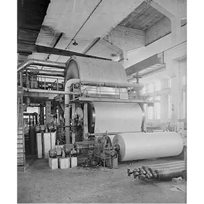A historical black-and-white photograph shows a large Yankee paper machine inside a factory. The machine has multiple rollers and is producing a wide sheet of paper wound into large rolls. Mechanical components and spare parts are visible on the floor, and the high-ceilinged space has windows providing natural light, typical of early 20th-century manufacturing facilities.
