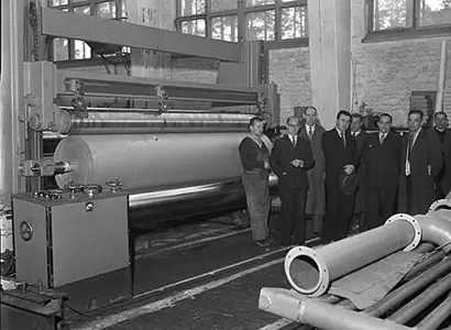 A black-and-white photograph depicts a group of men, some in suits and others in work attire, standing beside a large industrial paper machine at Rautpohja, with massive rollers inside a factory setting. The machine appears to be producing or processing a wide roll of paper, and additional large cylindrical components are visible in the foreground, indicating heavy manufacturing equipment.