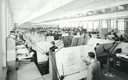 A historical black-and-white photograph shows a large drafting office filled with rows of drafting tables and individuals working on technical drawings. The space is well-lit with tall windows and overhead lighting, and the workers appear focused on detailed architectural or engineering projects.
