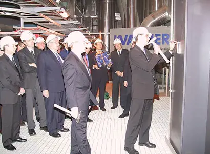 A group of people dressed in formal business attire and wearing white safety helmets are gathered inside an industrial facility. One individual in the foreground is signing or writing on a large metallic surface, while others look on. The scene suggests a ceremonial or official event, related to the inauguration of industrial equipment.