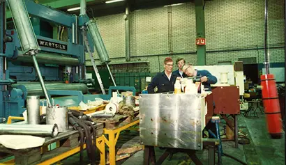 Järvenpää workshop in 1983. The image shows three men working together in an industrial workshop, surrounded by heavy machinery, tools, and equipment parts. A large blue machine labeled “Wärtsilä” is visible on the left side, indicating the manufacturer. The workers appear to be inspecting or assembling components on a metal workbench in a spacious factory setting.