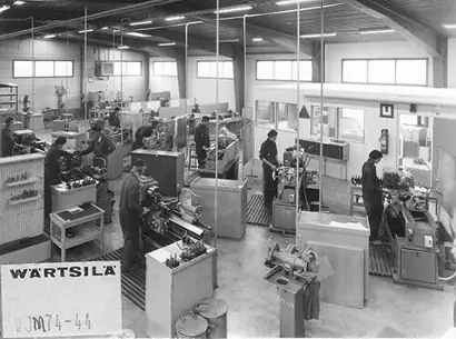 A historical black-and-white photograph shows a machine workshop with multiple workers operating lathes and other metalworking machines. The large, organized space has high windows for natural light and partitioned sections, suggesting a structured manufacturing environment. A sign reading “Wärtsilä” is visible in the foreground, indicating the company's association with the facility.