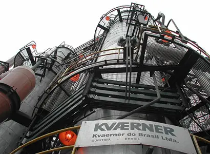 An upward view of large industrial towers and metal structures, featuring interconnected pipes, platforms, and railings. The foreground shows a sign reading “Kværner do Brasil Ltda., Curitiba – Brasil,” indicating the facility’s operator. The overall scene suggests a heavy industrial plant, likely involved in chemical, energy, or processing operations.