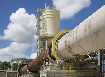 A large industrial rotary kiln system is shown outdoors under a bright blue sky with scattered clouds. The structure consists of a massive cylindrical drum supported by a metal framework and connected to a tall beige silo with ladders and platforms. The equipment is part of a heavy manufacturing or processing facility, likely used for material production or thermal treatment.