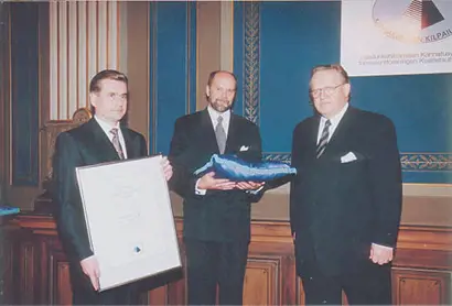 Three men, Martti Ahtisaari, Hannu Mälkiä and Martti Karttunen, in formal business attire are participating in an award ceremony inside an elegant room with ornate wooden paneling and blue walls. One man is holding a large framed certificate, while another is presenting an object wrapped in blue fabric. A banner with a logo is partially visible in the background, indicating an official or professional recognition event.