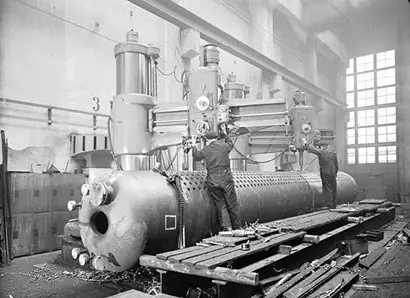 A historical black-and-white photograph shows two Tampella mechanics operating a large industrial drilling machine on a cylindrical metal component, likely a boiler or pressure vessel. The workshop is spacious with high ceilings, tall windows providing natural light, and metal shavings scattered on the floor, indicating active heavy manufacturing or machining work.