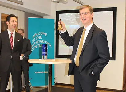 A man in a business suit is holding a small bell in one hand, standing beside a round table. Another man in a suit stands nearby, and a blue banner in the background reads “Welcome to the NASDAQ OMX”. The setting appears to be a formal financial or corporate event, such as a market opening ceremony.