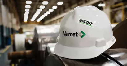 A white safety helmet with the logos "Valmet" and "Beloit" printed on it is placed on top of large industrial machinery or equipment inside a manufacturing facility. The background shows a factory setting with bright overhead lights and blurred industrial components, indicating an operational production environment.