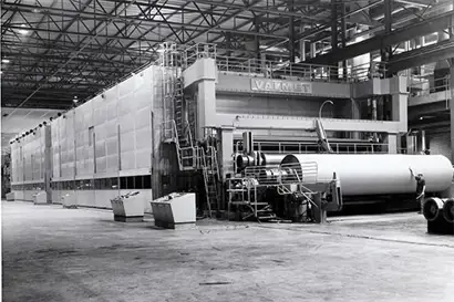 A black-and-white photograph depicts a massive industrial paper machine inside a spacious factory. The large structure features multiple rollers and control panels on the side, with a huge roll of finished paper being processed. The word “Valmet” is prominently visible on the upper section of the machine, indicating the manufacturer.
