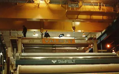 The image shows a large industrial setting with heavy machinery and overhead cranes. A prominent orange crane marked "VALMET" is visible at the top, while large rollers and components labeled "TAMFELT" occupy the lower part of the scene. Several workers are standing on the overhead structure, indicating maintenance or installation activities in a paper or material processing facility.