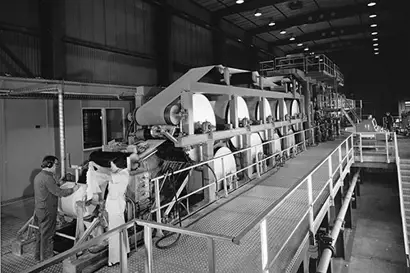 A black-and-white photograph shows a large industrial pilot machine with multiple rollers in operation. A worker wearing protective gear is handling the material at the beginning or end of the machine, which is set within a spacious, high-ceilinged factory environment. Walkways with railings run alongside the machine for operational access.