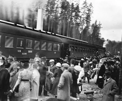 A historical black-and-white photograph depicts a large crowd of people gathered near a stationary passenger train. The scene shows men and women dressed in early-to-mid 20th-century attire, including hats and coats, suggesting a significant public event or travel occasion. Tall trees line the background, indicating a rural or forested setting. Located in Rautpohja, 1938.