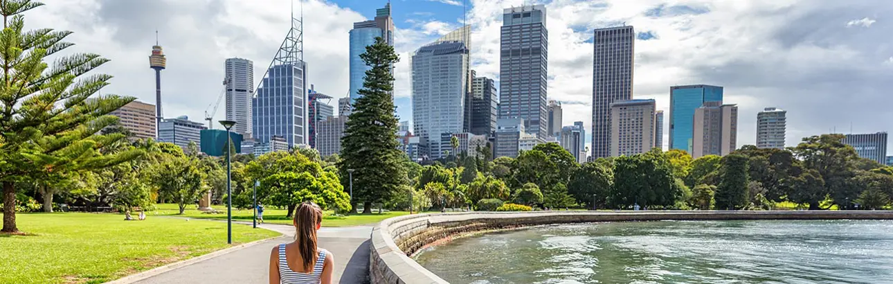 A person is walking along a curved waterfront path in a green park, with a city skyline featuring modern skyscrapers in the background. The scene is bright and sunny, with blue skies and scattered clouds. Trees and grassy areas frame the view, creating a contrast between nature and urban architecture.