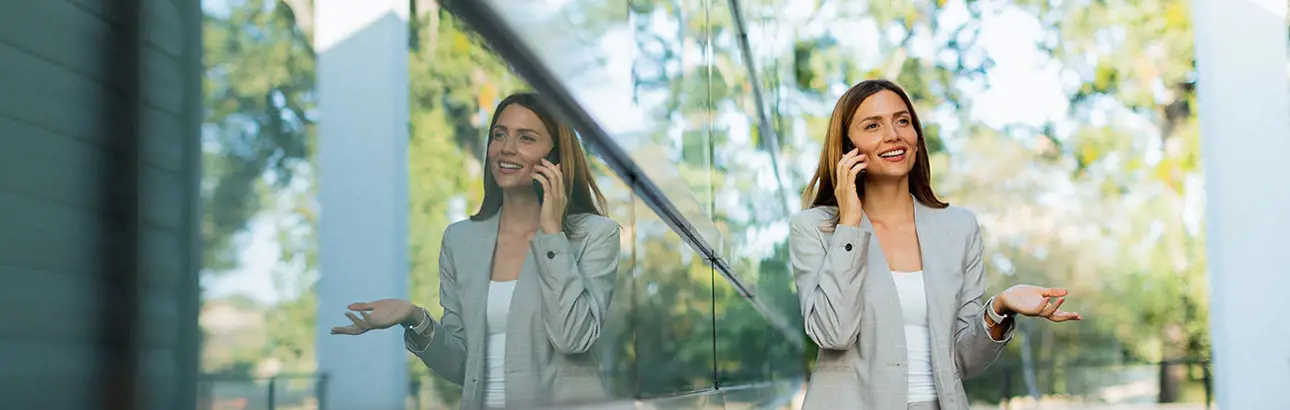 A woman in a light gray blazer and white top is standing outdoors, talking on the phone with one hand holding the phone and the other hand raised in a questioning or expressive gesture. She is next to a reflective glass wall, which creates a clear mirror image of her. The background shows blurred green trees, suggesting an outdoor or garden setting.