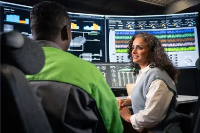 Two people are seated in front of multiple large screens displaying complex data visualizations, charts, and system controls. One person wears a green and gray work jacket, while the other is dressed in a white shirt with a light blue vest. They appear to be discussing or monitoring industrial processes in a high-tech control room environment.
