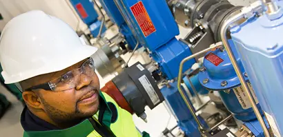 A Valmet worker wearing a white safety helmet and a high-visibility jacket is inspecting or operating a section of industrial piping and machinery. The equipment features multiple blue valves, metal pipes, and control elements, indicating a controlled processing or manufacturing environment.