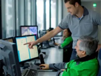 Two people are in a control room with multiple computer monitors displaying system data. One person seated, wearing a green jacket, is operating the computer, while another person standing beside them is pointing at the screen, likely giving instructions or explaining something. Other operators can be seen working in the background.