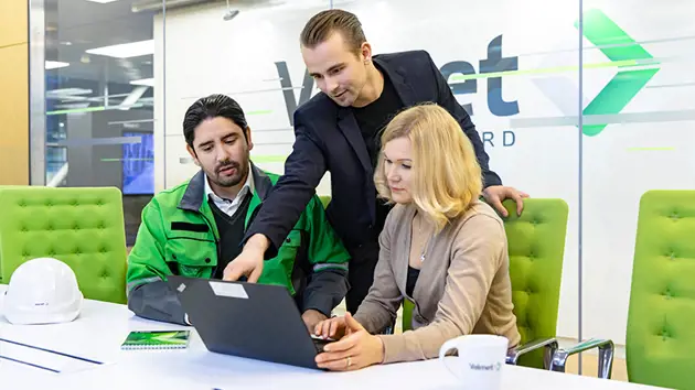 Three people gathered around a computer; one person is standing and pointing at the screen while the other two are seated on either side.