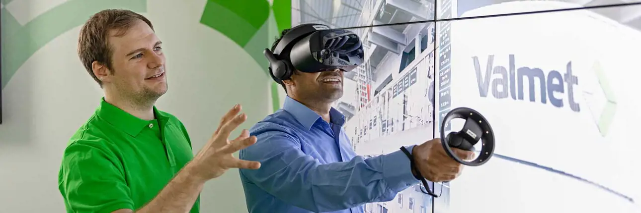 Two people standing side by side; one is wearing VR glasses and holding a remote, while the other appears to be guiding or assisting.