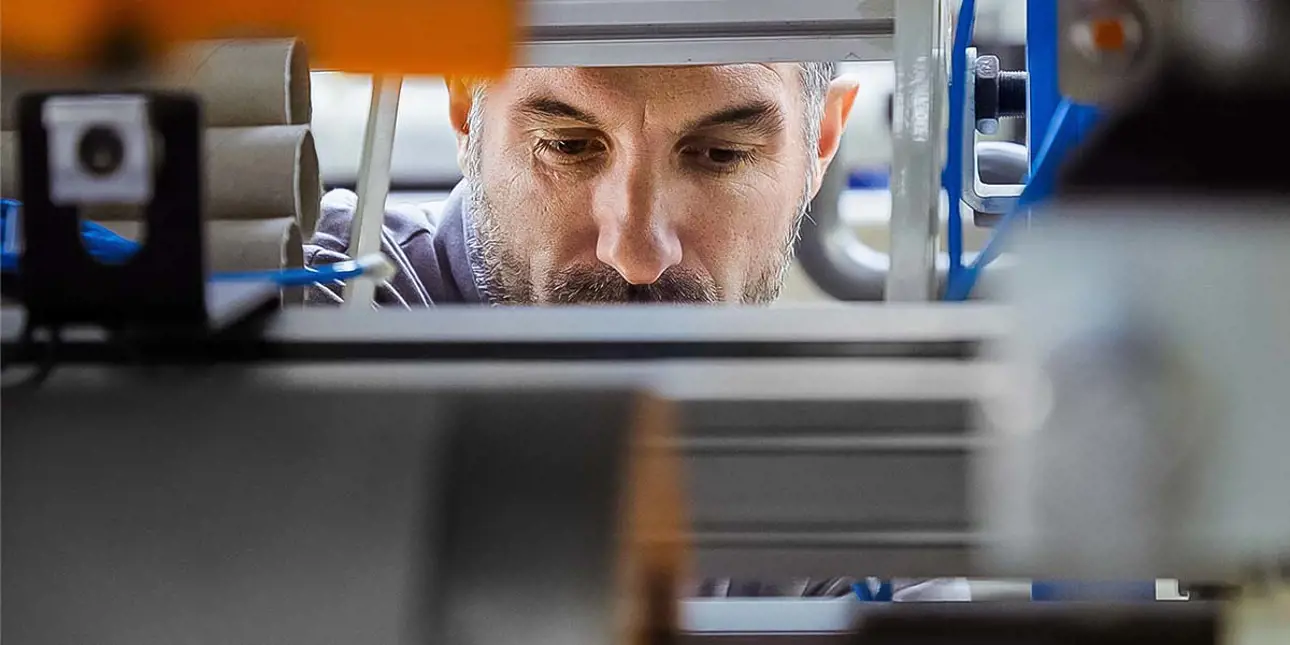 A technician carefully inspecting machinery components through a narrow opening, with focus on his concentrated expression and the industrial setup surrounding him.