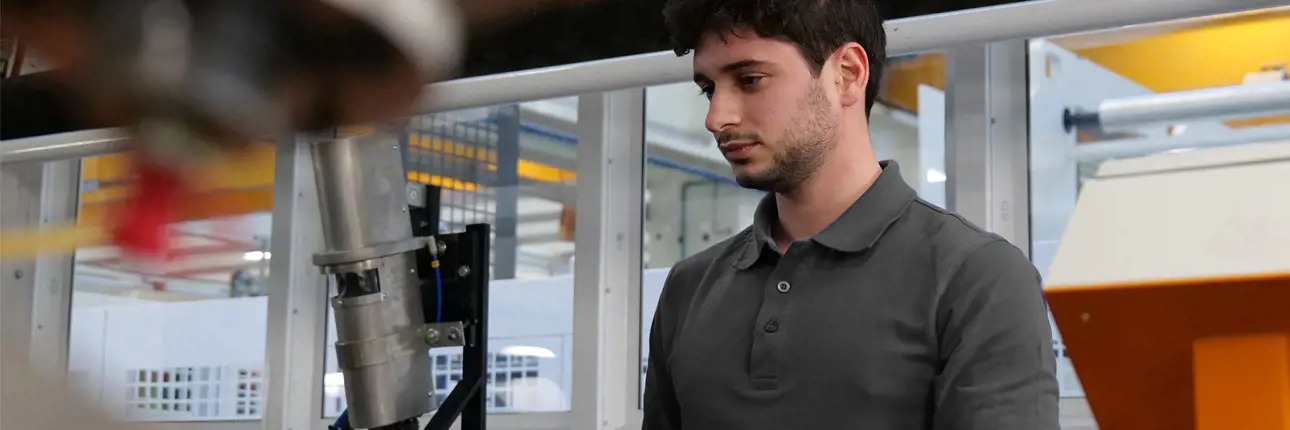 A technician in a dark grey polo shirt closely observing machinery inside an industrial facility.