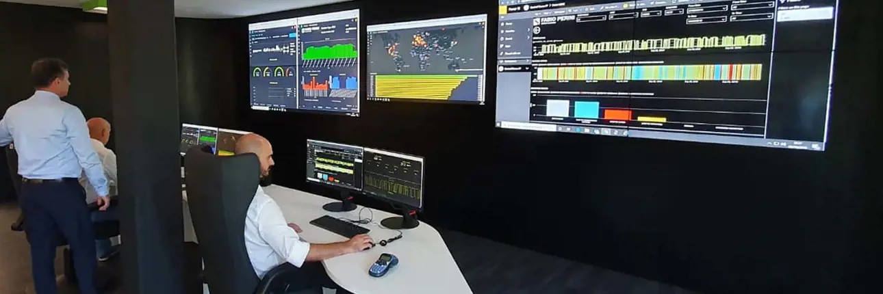 Several people monitoring real-time industrial data on multiple screens in a control room, with dashboards and performance metrics displayed on large wall-mounted monitors.