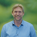 a smiling man in a blue shirt standing infront of a green studio screen